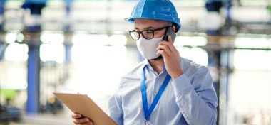 Endüstriyel fabrikada çalışan koruyucu maskeli ve telefonlu teknisyen veya mühendis.