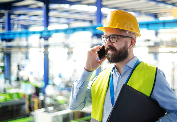 Technician or engineer with hard hat standing in industrial factory, using telephone.