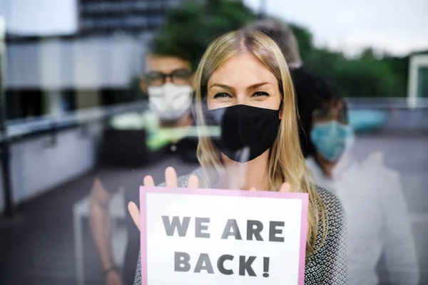 Woman with face mask back at work in office after lockdown, holding we are back sign.