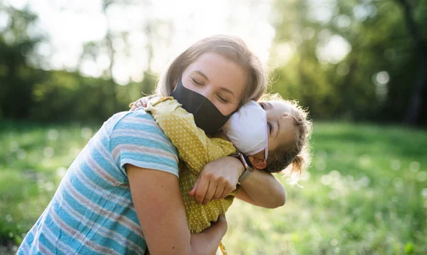 Vista lateral de la madre feliz abrazando a la pequeña hija en la naturaleza, con máscaras faciales . —  Fotos de Stock
