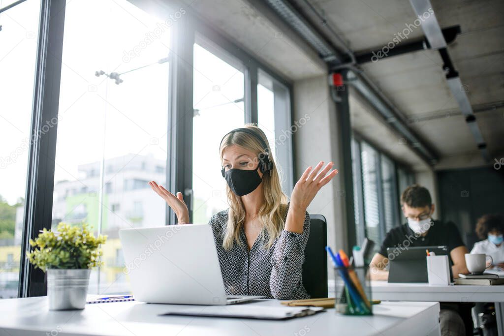 Young woman with face mask back at work in office after lockdown, having video call.