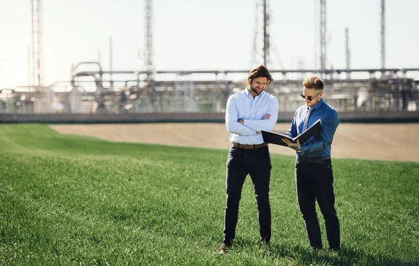 Two young engineers standing outdoors by oil refinery, discussing issues. — Stock Photo, Image