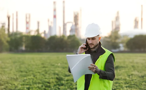 Young engineer with smartphone standing outdoors by oil refinery. Copy space. — Stock Photo, Image