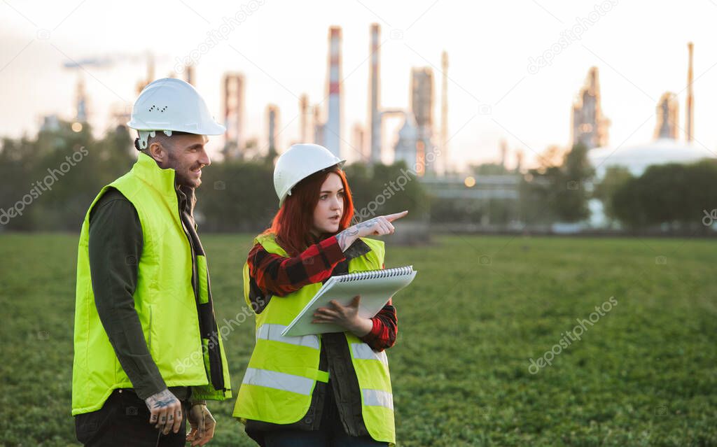 Two young engineers standing outdoors by oil refinery, discussing issues.