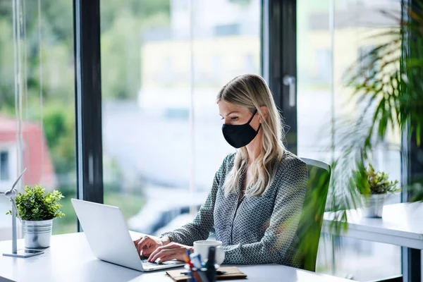 Jovem com máscara facial de volta ao trabalho no escritório após o bloqueio, usando laptop . — Fotografia de Stock