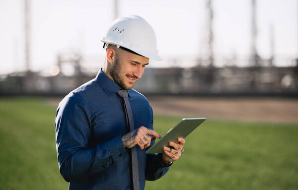Young engineer with hard hat and tablet standing outdoors by oil refinery.