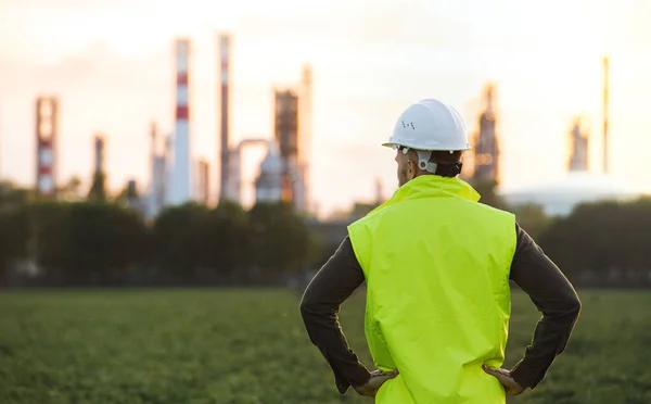 Rear view of young engineer standing outdoors by oil refinery, arms on hips. — Stock Photo, Image