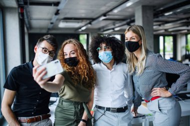 Young people with face masks back at work in office after lockdown, taking selfie. clipart