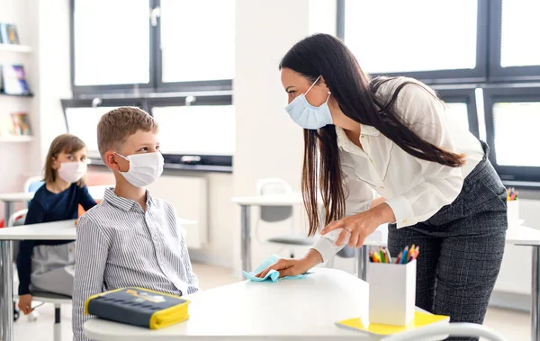 Maestra y niños con mascarilla en la escuela, desinfectando escritorios . — Foto de Stock