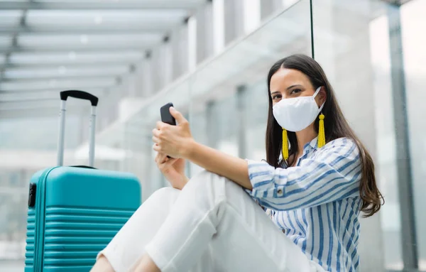 Mujer joven con teléfono inteligente va de vacaciones, con máscaras faciales en el aeropuerto . — Foto de Stock
