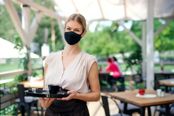 Официантка в маске для лица обслуживает клиентов на открытом воздухе на террасе ресторана . — стоковое фото