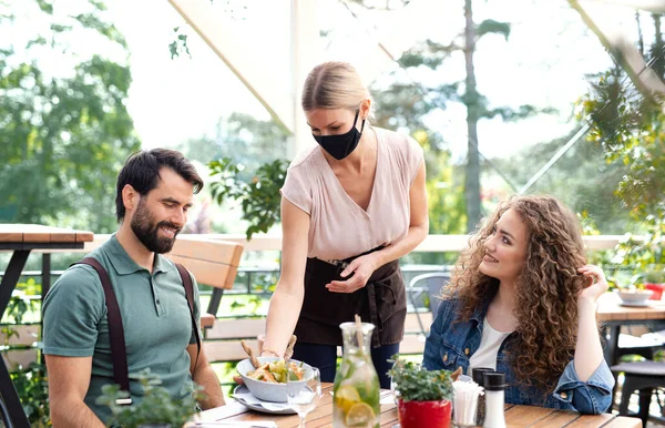 Waitress with face mask serving happy couple outdoors on terrace restaurant. — Stock Photo, Image