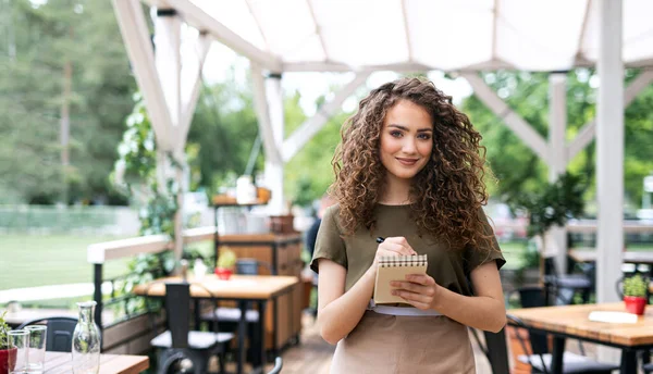 Waitress with order pad standing outdoors on terrace restaurant, looking at camera. — Stock Photo, Image