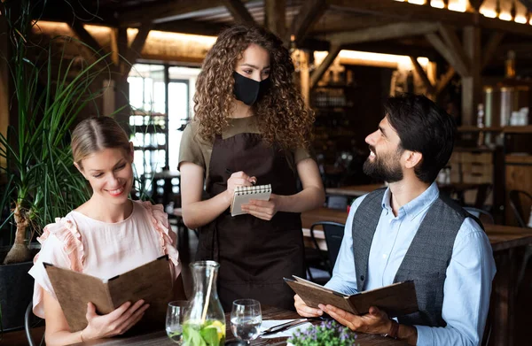 Camarera con mascarilla que sirve feliz pareja en el interior del restaurante . — Foto de Stock