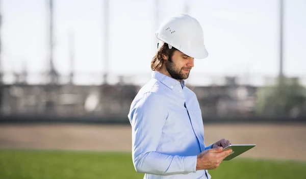 Ingeniero joven con el sombrero duro parado al aire libre por la refinería de petróleo, usando la tableta . —  Fotos de Stock