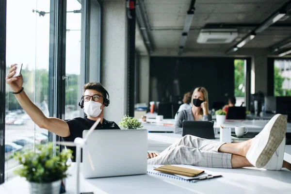 Young man with face mask back at work or school in office after lockdown, taking selfie. — Stock Photo, Image