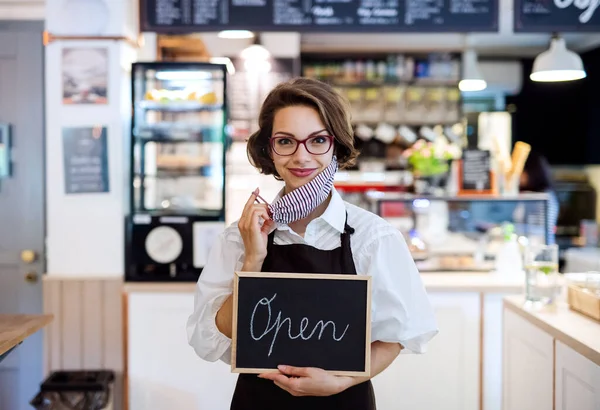 Νεαρή γυναίκα με μάσκα προσώπου που εργάζεται σε καφετέρια, κρατώντας ανοιχτή πινακίδα. — Φωτογραφία Αρχείου