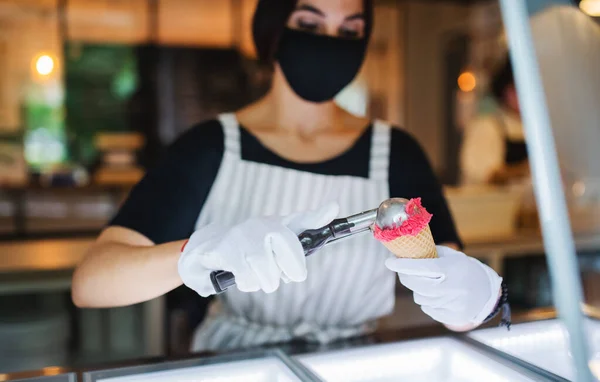 Giovane donna con maschera viso che lavora al chiuso in caffetteria, servendo gelato. — Foto Stock