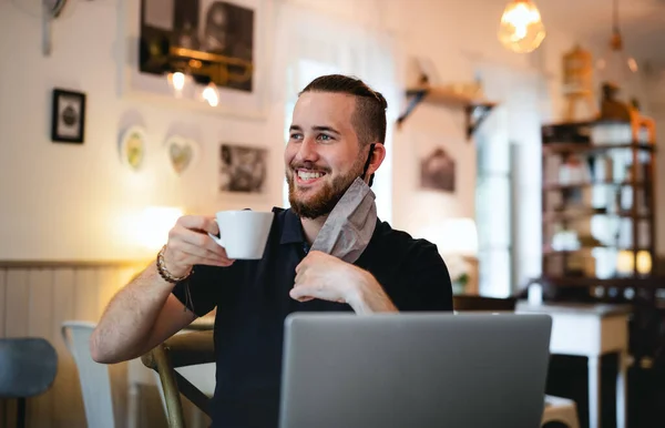 Young man with face mask and laptop indoors in cafe, drinking coffee. — Stock Photo, Image