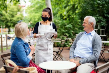 Yüzü maskeli genç bir garson kafede çalışıyor, kıdemli müşterilere hizmet ediyor..