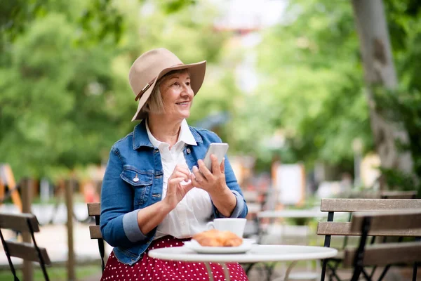 Senior woman with hat sitting outdoors in cafe, using smartphone. — Stock Photo, Image