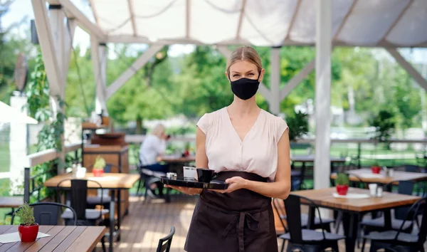 Официантка в маске для лица обслуживает клиентов на открытом воздухе на террасе ресторана . — стоковое фото