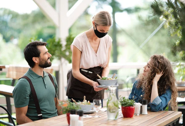 Waitress with face mask serving happy couple outdoors on terrace restaurant. — Stock Photo, Image