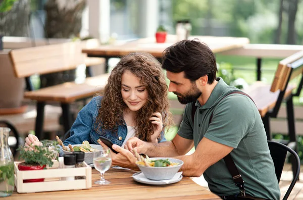 Joyeux couple assis à l'extérieur sur la terrasse restaurant, en utilisant un smartphone. — Photo