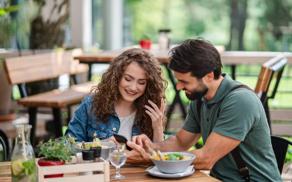 Happy couple sitting outdoors on terrace restaurant, using smartphone. — Stock Photo, Image