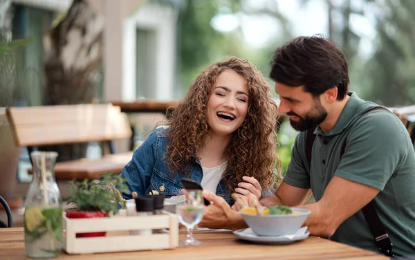 Šťastný pár sedí venku na terase restaurace, pomocí smartphone. — Stock fotografie