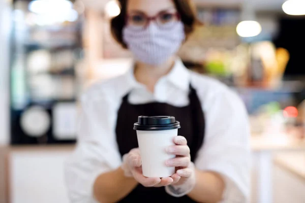 Giovane donna con maschera viso che lavora al chiuso in caffetteria, tenendo il caffè. — Foto Stock