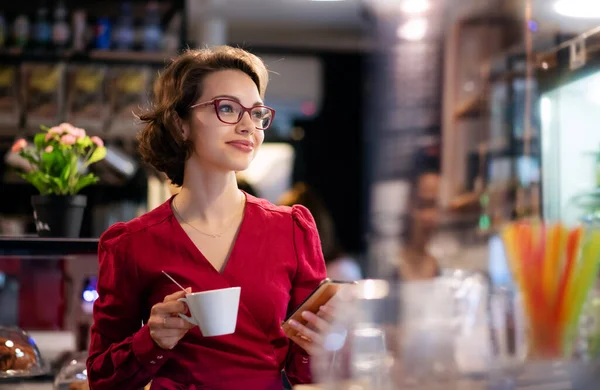Νεαρή γυναίκα με καφέ σε εσωτερικό χώρο στο cafe, χρησιμοποιώντας smartphone. — Φωτογραφία Αρχείου