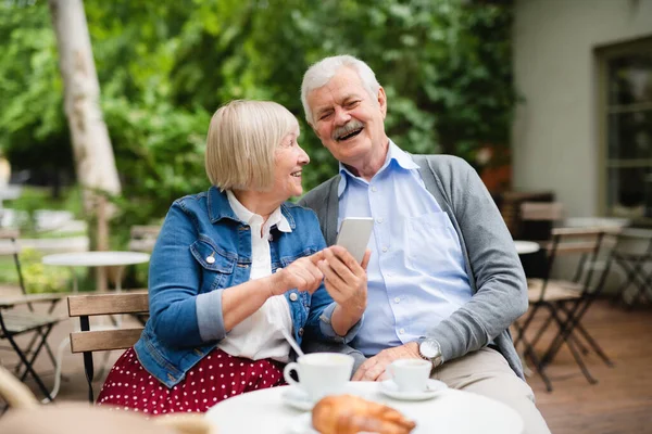 Glückliches verliebtes Senioren-Paar sitzt draußen im Café und nutzt Smartphone. — Stockfoto