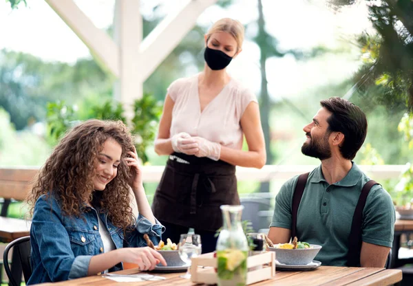 Kelnerka z maską twarzy serwująca szczęśliwą parę na świeżym powietrzu na tarasie restauracji. — Zdjęcie stockowe