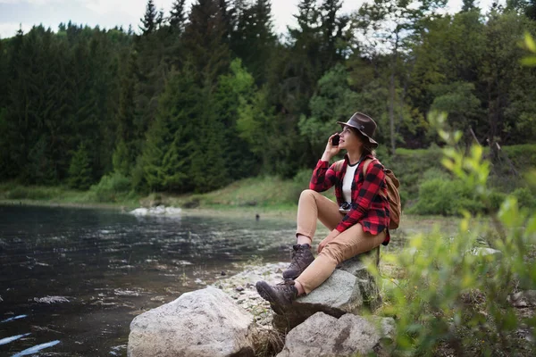 Jonge vrouw zit aan het meer op een wandeling buiten in de zomer natuur, met behulp van telefoon. — Stockfoto
