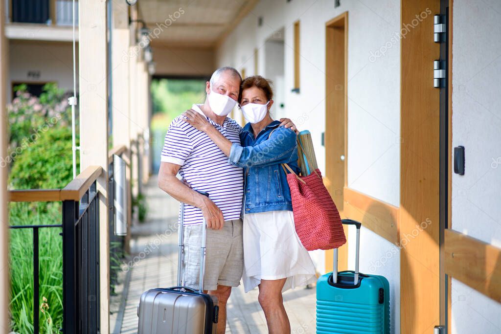 Senior couple with face masks and luggage outside apartment on holiday.