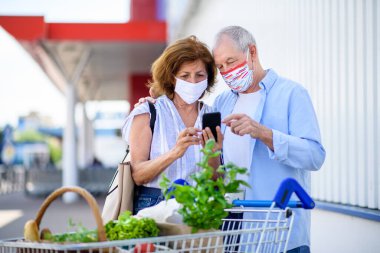 Şehirdeki süpermarketin dışında maskeli ve akıllı telefonlu yaşlı çift..