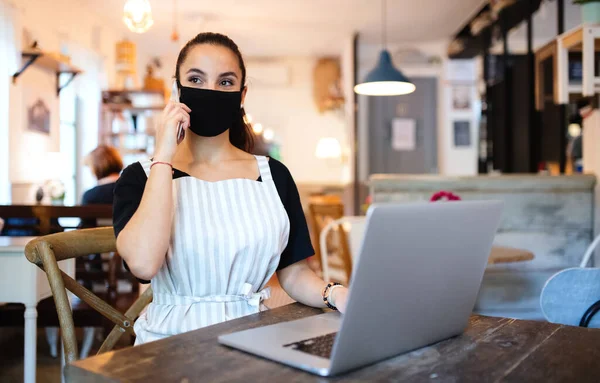 Jovem com avental, smartphone e laptop trabalhando no café . — Fotografia de Stock