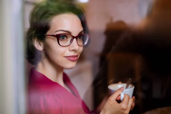 Jonge vrouw met koffie binnen in café. Door glas geschoten.. — Stockfoto