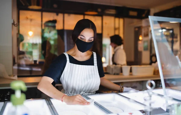 Νεαρή γυναίκα με μάσκα προσώπου που εργάζεται σε εσωτερικούς χώρους σε καφετέρια, απολυμαίνοντας τις επιφάνειες. — Φωτογραφία Αρχείου