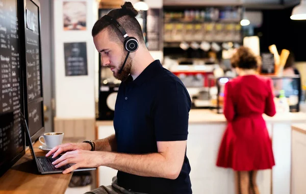 Jeune homme avec écouteurs et ordinateur portable à l'intérieur dans le café, travaillant. — Photo