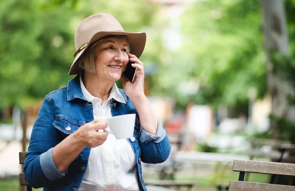Femme âgée avec chapeau assis à l'extérieur dans un café, en utilisant un smartphone. — Photo