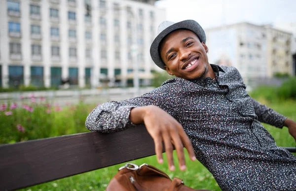 Веселый молодой чернокожий мужчина сидит на скамейке на открытом воздухе в городе. — стоковое фото