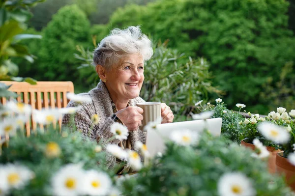 夏天，一位拿着笔记本电脑和咖啡坐在阳台上休息的老年妇女. — 图库照片