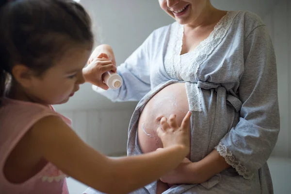 Kleines Mädchen cremt Bauch einer schwangeren Mutter im Bad zu Hause ein. — Stockfoto