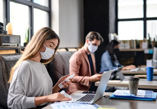 Retrato de jóvenes empresarios con máscaras faciales trabajando en interiores en oficina, desinfectando laptop. — Foto de Stock