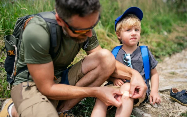 Father with small son hiking outdoors in summer nature, putting plaster on knee. — Stock Photo, Image