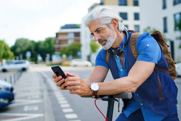 Dojrzały człowiek dojeżdżający do pracy z hulajnogą elektryczną na świeżym powietrzu w mieście, za pomocą smartfona. — Zdjęcie stockowe