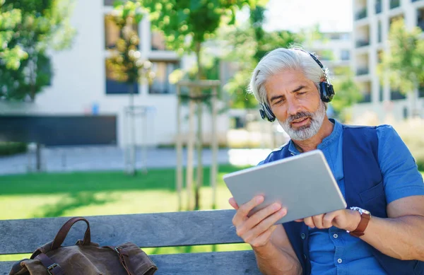 Porträt eines reifen Mannes mit Kopfhörern, der draußen in der Stadt sitzt und ein Tablet benutzt. — Stockfoto