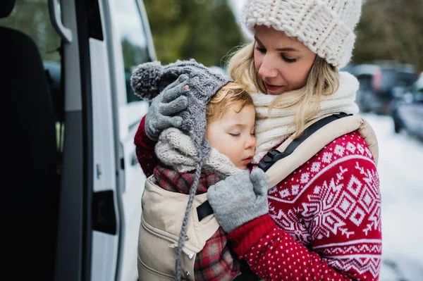 Mor med sovande liten dotter i bärstol stående med bil i vinternaturen. — Stockfoto